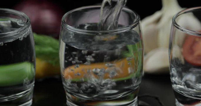 Pour alcohol vodka from a bottle in two shot glass. Background with vegetables