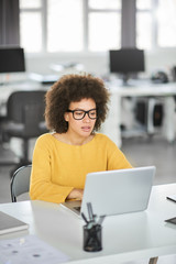 Serious mixed race businesswoman dressed casual sitting in modern office and using laptop.