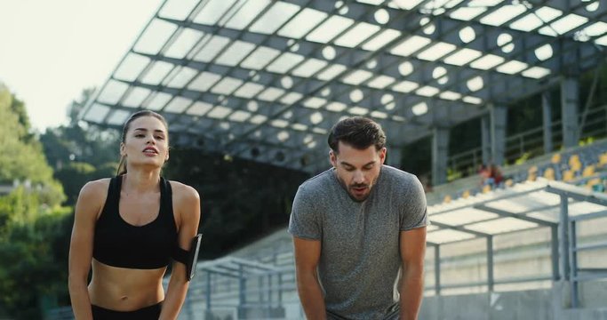 Young Caucasian sporty woman and man jogging at the stadium and stopping to have a rest for a minute, then smiling to the camera. Summer sunny day.
