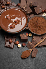 glass bowl of chocolate cream or melted chocolate, pieces of chocolate and hazelnuts on dark concrete background