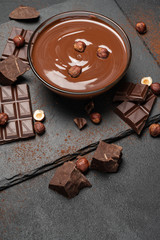 glass bowl of chocolate cream or melted chocolate, pieces of chocolate and hazelnuts on dark concrete background