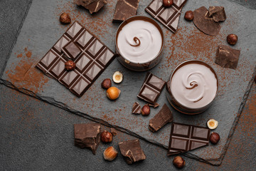 glass bowl of chocolate cream or melted chocolate, pieces of chocolate and hazelnuts on dark concrete background