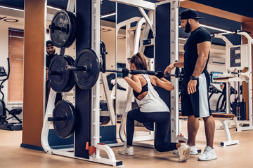 Attractive young woman does barbell squats with trainer in gym