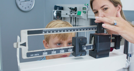 Doctor weighing little boy patient on scale in exam room. Female pediatrician and child performing...