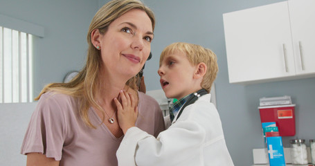 Obraz premium Cute little boy pretending to be a doctor with his mother while in pediatricians office. Child wearing oversized lab coat and stethoscope using otoscope