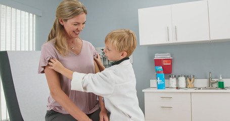 Cute little boy pretending to be a doctor with his mother while in pediatricians office. Child wearing oversized lab coat and stethoscope using otoscope