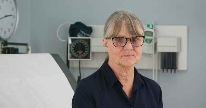 Senior woman patient sitting in medical exam room waiting for the doctor turns to look at the camera. Older female in clinic or hospital waiting for her primary care physician