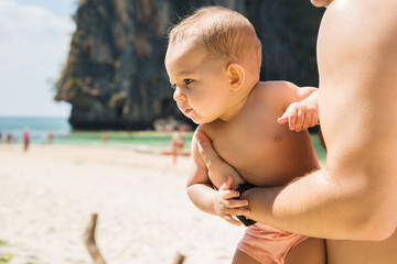 A small infant baby in the arms of dad, very much interested, turned away. Reaches for something, looing for mom. Toddler on sunny tropical beach.