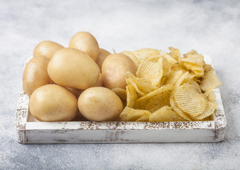 Fresh organic homemade potato crisps chips with raw yellow potatoes in white wooden box on light kitchen table background.