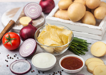 Fresh organic homemade potato crisps chips in glass bowl with sour cream and red onions on light kitchen table background. Box of fresh yellow potatoes, bowl of ketchup and green onion and tomatoes