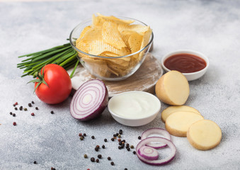 Fresh organic homemade potato crisps chips in glass bowl with sour cream and red onions on light kitchen table background. Tomato with ketchup and green onion