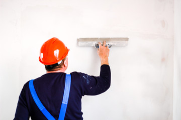 A builder in an orange construction cup in his hands with a spatula and a white putty. Blue form...