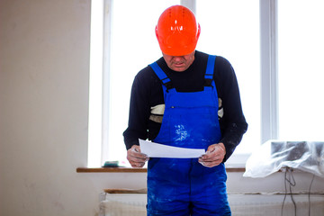 A builder in an orange construction catcher in his hands with drawings. Blue form combining. Repair in the apartment.