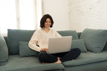 Fototapeta premium Happy young woman working on her laptop sitting cross-legged on the sofa. In technology concept