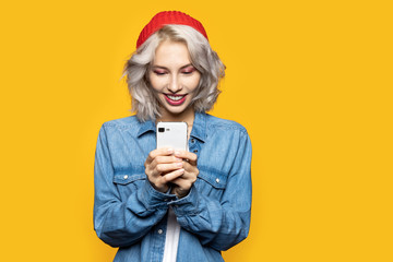Portrait of charming blonde girl texting with someone on smartphone. Fashionably dressed lady in jacket. Communication and lifestyle concept. Isolated on yellow background - obrazy, fototapety, plakaty