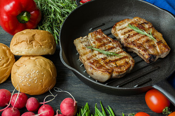 Grilled steak on a round grill pan, garnished with spices for meat, rosemary, greens and vegetables on a dark wooden background.