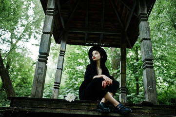 Sensual girl all in black, red lips and hat. Goth dramatic woman sitting on wooden arch at forest.