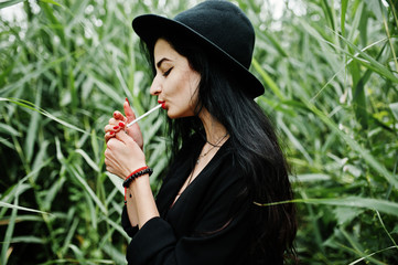 Sensual smoker girl all in black, red lips and hat. Goth dramatic woman smoking on common reed.