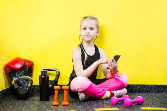 Young child girl with a mobile phone sitting on the floor near the dumbbells, boxing gloves and a bottle of water on the floor. Ready to workout at the gym. Sport and healthy concept