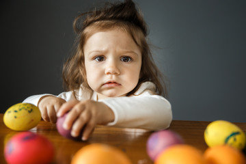 Fototapeta na wymiar Upset baby girl is sitting behind the table with a bunch of colored eggs laying on it. She is looking at the camera while holding an egg with her left hand. Isolated on gray background.