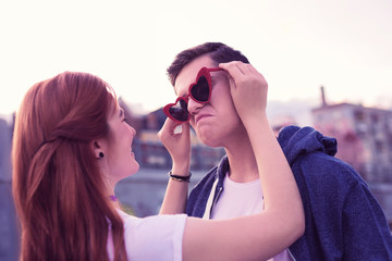 Laughing ginger girl attaching funky red sunglasses to the face of her boyfriend
