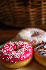 White, pink and brown glazed donuts on wooden background and near rattan basket .