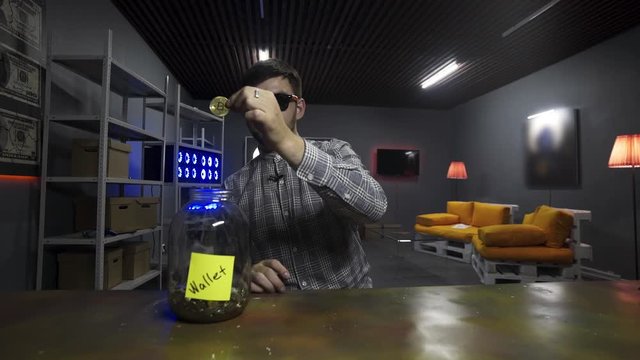 Nice bearded smiling man wearing sunglasses and checkered shirt, holds big gold bitcoin and drops it into huge glass jar with other coing in front of camera in room with TV on wall and yellow sofa