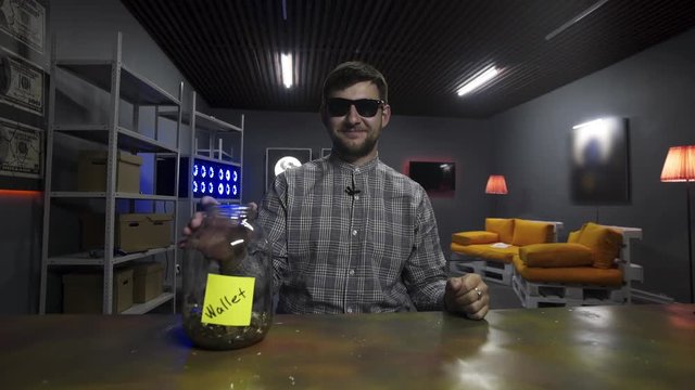 Funny bearded young man wearing sunglasses and checkered shirt, holds big gold bitcoin and drops it into huge glass jar with other coing in front of camera in room with TV on wall and yellow sofa