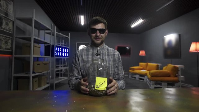 Smiling bearded young man wearing sunglasses and checkered shirt, holds gold bitcoin and drops it into huge glass jar with other coing in front of camera in room with TV on wall and yellow sofa