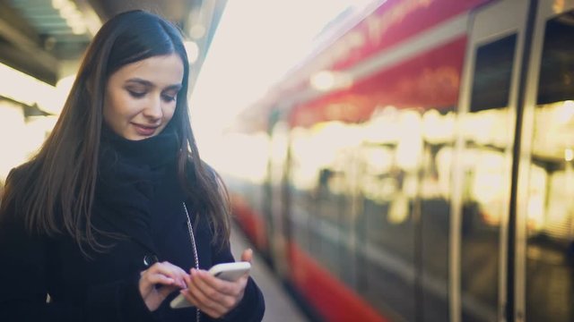 Young lady typing on smartphone on platform near train and smiling to camera