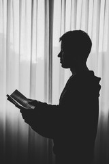 Side view of silhouette boy standing by window reading a book