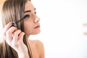 Obraz premium Kobieta nakładająca czarny tusz do rzęs na rzęsy pędzlem do makijażu. Młoda piękna kobieta stosując makijaż tusz do rzęs na oczy w łazience. Piękna brunetka dziewczyna nakładanie makijażu na oczy w godzinach porannych.