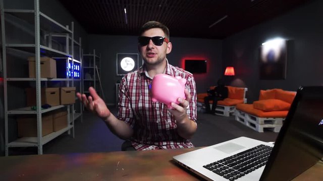 Cute bearded young man vlogger wearing sunglasses and red checkered shirt, emotionally speaks at camera holding pink piggy bank in hand, while someone in black clothes sits on orange sofa with laptop.