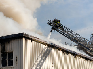  Fireman fighting a building fire at the top of an extended ladder. A hose attached to the ladder pours water through the smoke. The fireman is wearing breathing apparatus. Room for text. 
