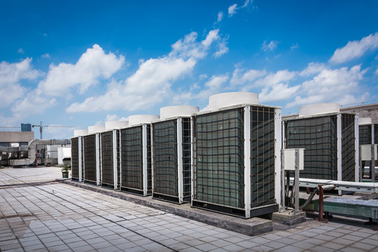 Square air-conditioning unit on the roof with a round fan. In the background gradually receding other units that are out of focus. On the right side light blue sky and commercial space.