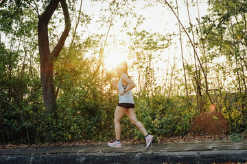 Young woman runner wears white sport vest, white cap, black short pant runs on the road to exercise in the evening while listens to music through earphone. Female runner runs on the city road.