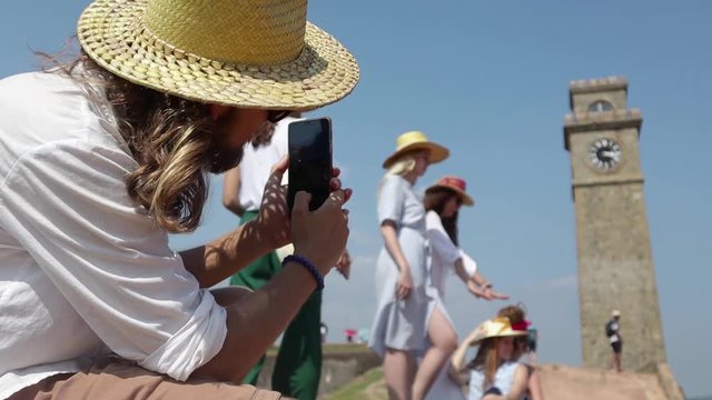 A young man in a straw hat taking a picture of a group of girls using his smartphone. Traveling with friends.