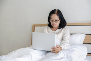 grandmother aged sitting on a bed in white bedroom with laptop in the morning time