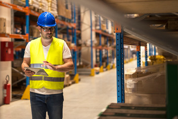 Warehouse worker checking inventory on digital tablet in large distribution warehouse storage area.