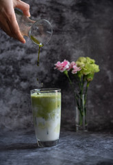 Hand of woman holding grass of green tea and pouring into a glass of milk with black concrete in background.
