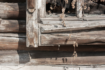 Fragment of a window without glass in the wall of an old log house close-up. Branch of dry hop on the window of an old house. Broken window with carved platband in the sun.