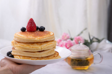 Front view of Pancakes on white plate topped with strawberry, rasberry and black currants and honey syrup and pink blooming flower in blackground.