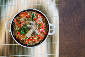 Salad with glass noodles, mushrooms and various vegetables in a plate on a bamboo Mat on a brown table. Top view, close-up, copy space