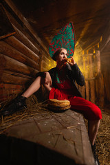 a girl with pancakes and caviar, in a red kokoshnik and black jacket, sits in a barn.