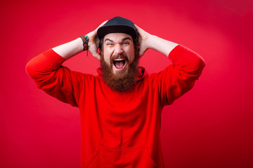 Handsome frustrated guy with beard screaming and putting hand on head, standing over red isolated ...