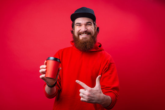 Happy smiling guy with beard in red hoody, pointing at take away cup over red wall
