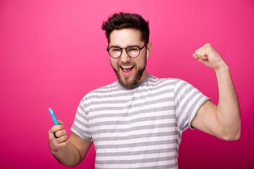 Portrait of a young man wearing glasses, and airpods, celebrate with rised arm while holding mobile