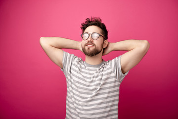 Happy young man wearing  glasses, holding hands behind head and looking up over pink background