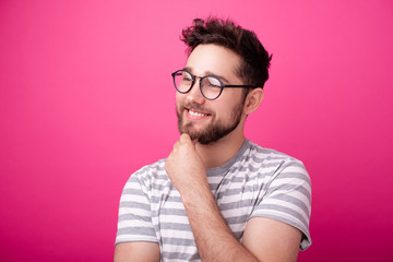 Fototapeta premium Handsome young bearded man wearing glasses, looking far away and touching his chin over pink background