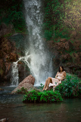 Fototapeta na wymiar Sexy woman posing on the rock among green tropical plants and beside beautiful waterfall with blue water. Fairy tale.